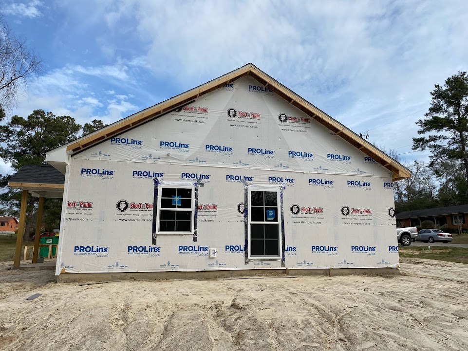 Wood-framed house under construction with plastic sheeting, exposed windows, blue permit sign, green garbage bin, bare dirt foreground, leafless tree, and clear blue sky