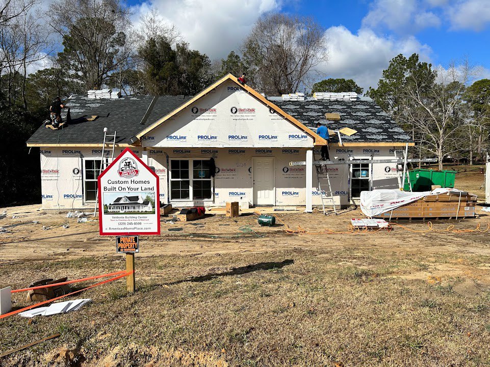 Partially built house with exposed framing, white window frame, stack of brown boxes on grass, large green tank beside structure, sign displaying house illustration, man