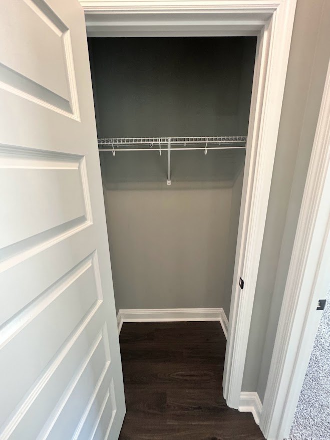 White closet with built-in shelf, dark wood flooring, white trim, and white door frame