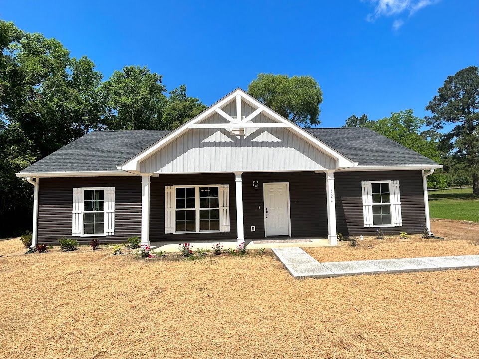 Two-story cottage-style home with white siding, white door with black handle, white-trimmed windows and shutters, front porch, grassy yard, mature trees, and Little White House