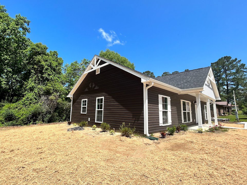 Two-story house with white trim, gray siding, and asphalt shingle roof, surrounded by a large grassy yard, mature trees, and blue sky with scattered clouds
