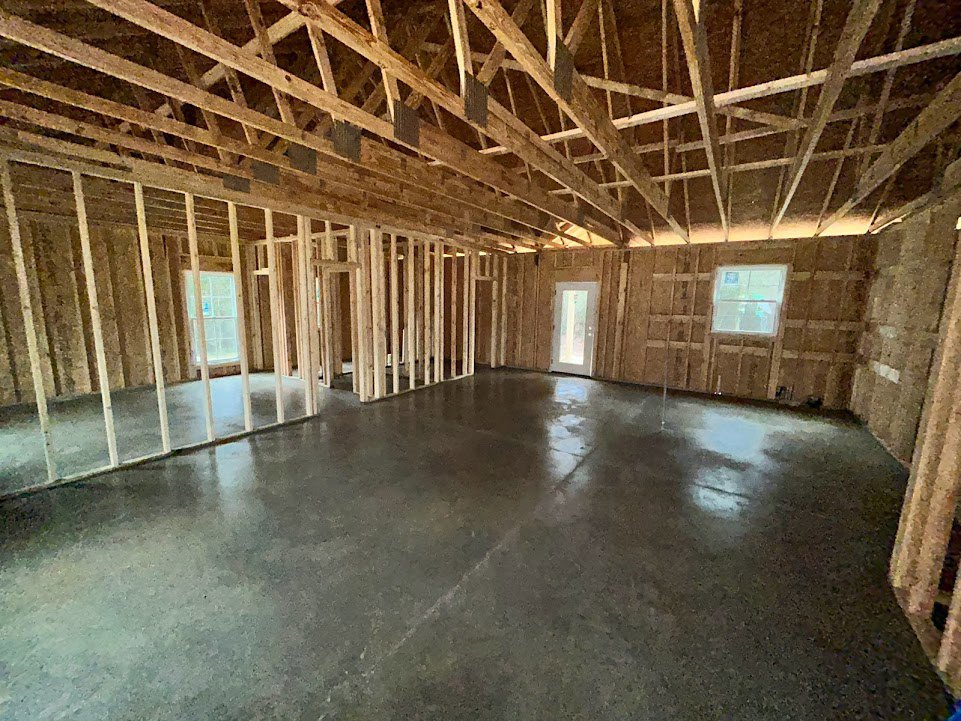Living room with exposed wood ceiling beams, large window letting in natural light, light-colored flooring, and partially finished walls with visible insulation.