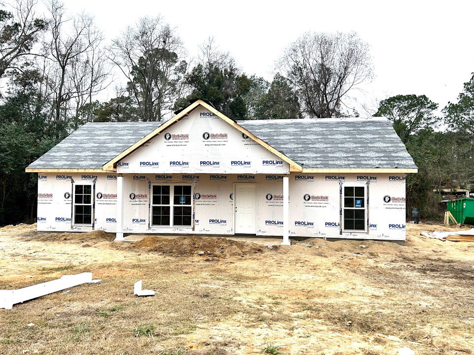 Partially built house with exposed roof, white door with black trim, multiple windows including one with a blue sign, surrounded by dirt and mature trees