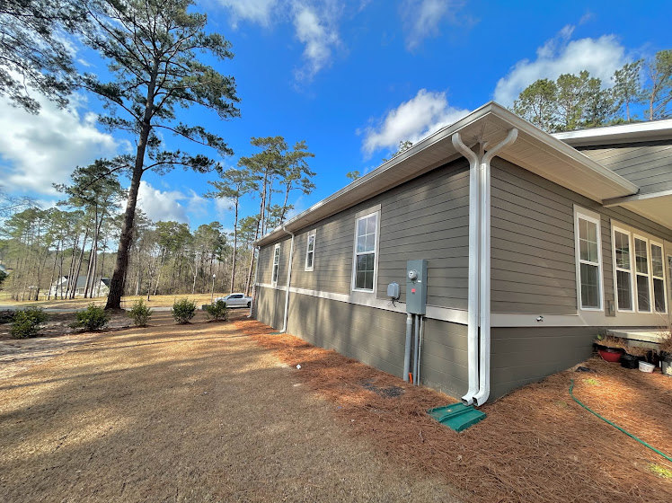 Two-story home with light siding, large windows, covered front porch, surrounded by mature trees and green lawn under partly cloudy sky