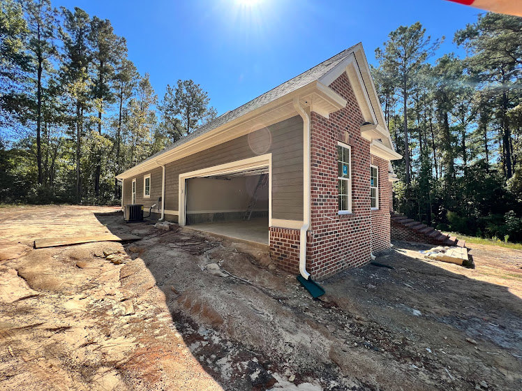 Framed house under construction with exposed wooden walls, surrounded by mature trees and open sky