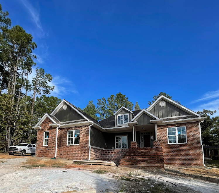 Two-story home under construction with exposed brick staircase, brick columns, and large window displaying a blue sign; surrounded by mature trees under a clear blue sky.