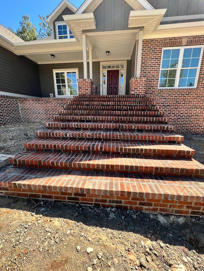 Brick staircase leading to red front door with white trim, adjacent window with white frame, pile of dirt beside brick wall, exterior porch area.