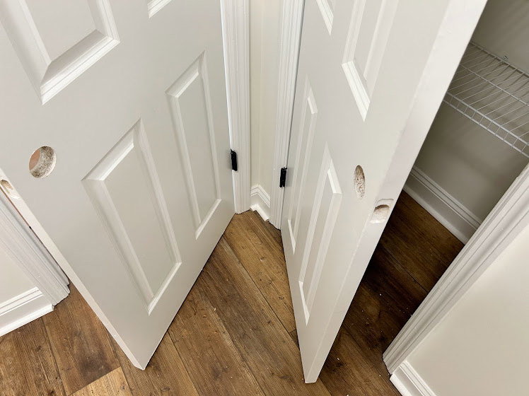 White wooden door with a round hole near the handle, surrounded by white wall molding and hardwood flooring