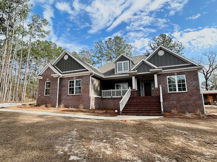 Red brick house with covered front porch, dirt yard, large windows, tree at side, and partly cloudy blue sky