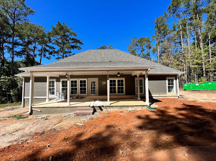 Wood-framed house under construction with exposed roof trusses, covered porch, white-framed windows, dirt and bricks on porch floor, surrounded by mature trees