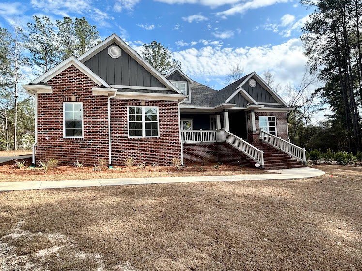 Two-story home with white siding, covered front porch, multiple windows, paved driveway alongside a dirt driveway, surrounded by trees under a partly cloudy sky