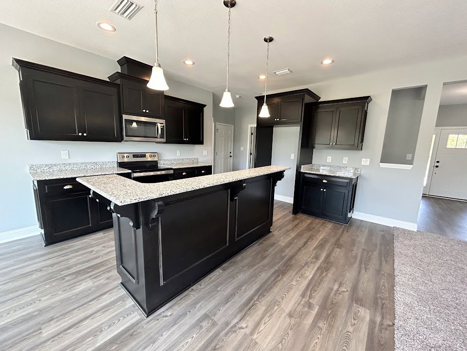 Spacious kitchen featuring a large central island with granite countertops, black cabinets with silver knobs and marble tops, stainless steel sink, light wood flooring, white door