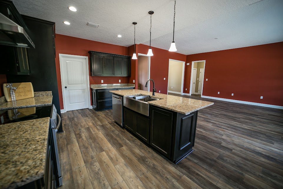 Kitchen with warm red walls, wood flooring, white cabinetry, black hardware, central island with sink, dishwasher integrated beside counter, white door with black handle