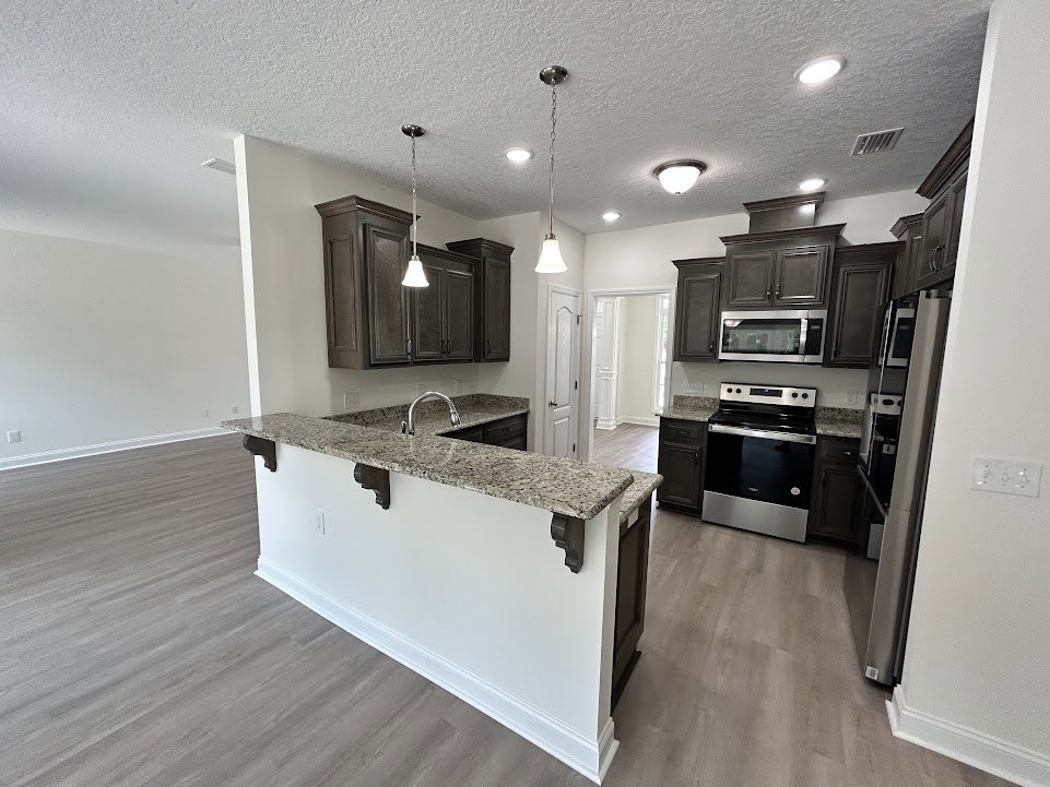 Granite kitchen island with wood flooring, black and silver stove, white cabinetry, stainless steel sink, metal bar on white wall, ceiling light fixture, window above counter