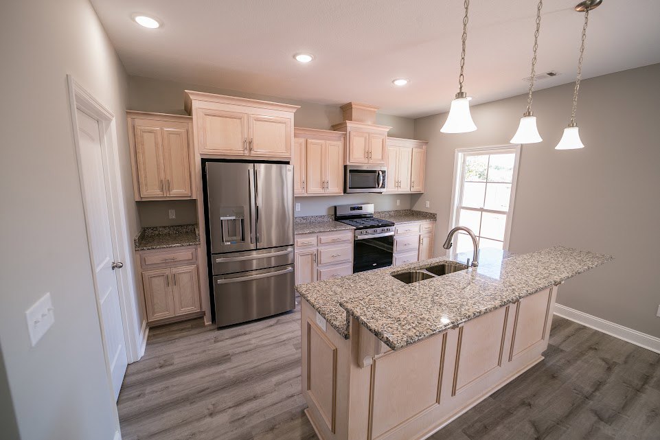 Kitchen with stainless steel refrigerator, wood flooring, white cabinetry, stone countertops, built-in stove and oven, undermount sink beneath window, and pendant lighting
