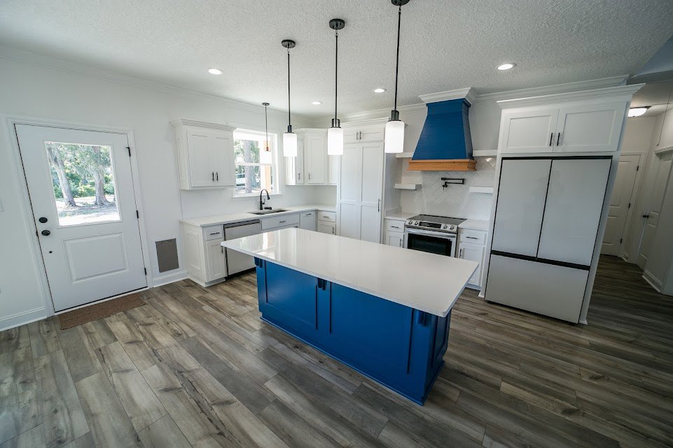 Blue kitchen island with white countertop, stainless steel stove, white refrigerator with black handles, blue vent hood mounted above wood shelf, white cabinetry, and white door