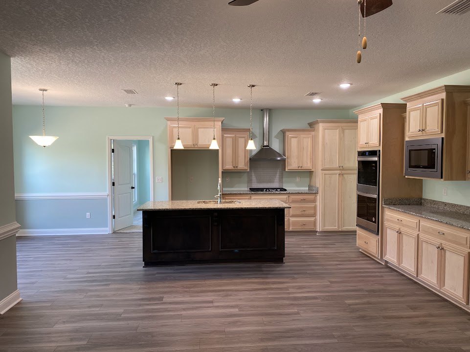 Open kitchen with wood flooring, central island with white countertop, built-in black microwave and double ovens on wall, stainless steel sink under window, recessed ceiling