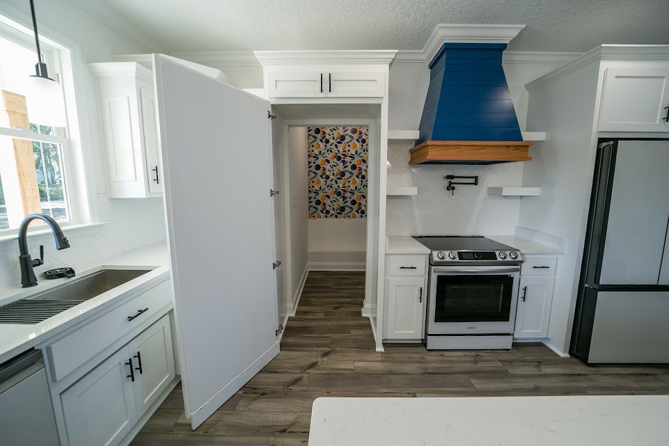 White stove with black door and matching refrigerator, blue vent hood above stove, black faucet over white tile floor, patterned oranges and leaves on backsplash, white cabinetry