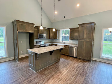 Kitchen with wide plank wood flooring, marble island countertop, brown wooden door, white walls, built-in cabinetry, stainless steel sink, and modern appliances.