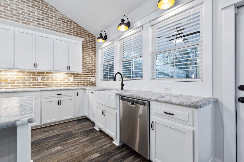 White shaker cabinets with black handles, stainless steel dishwasher and refrigerator, black faucet over a white sink, light fixture with yellow shade on ceiling, light gray