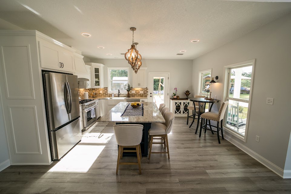 Modern kitchen with wood dining table and white cushioned chairs, stainless steel refrigerator, oven with towel draped on handle, light walls, and hardwood flooring