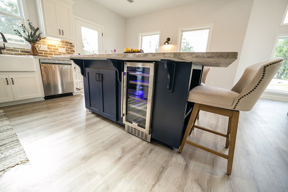 Open-concept kitchen featuring a wood bar with seating, stainless steel refrigerator and dishwasher, white cabinetry, stone countertops, a glass door illuminated by blue light, and