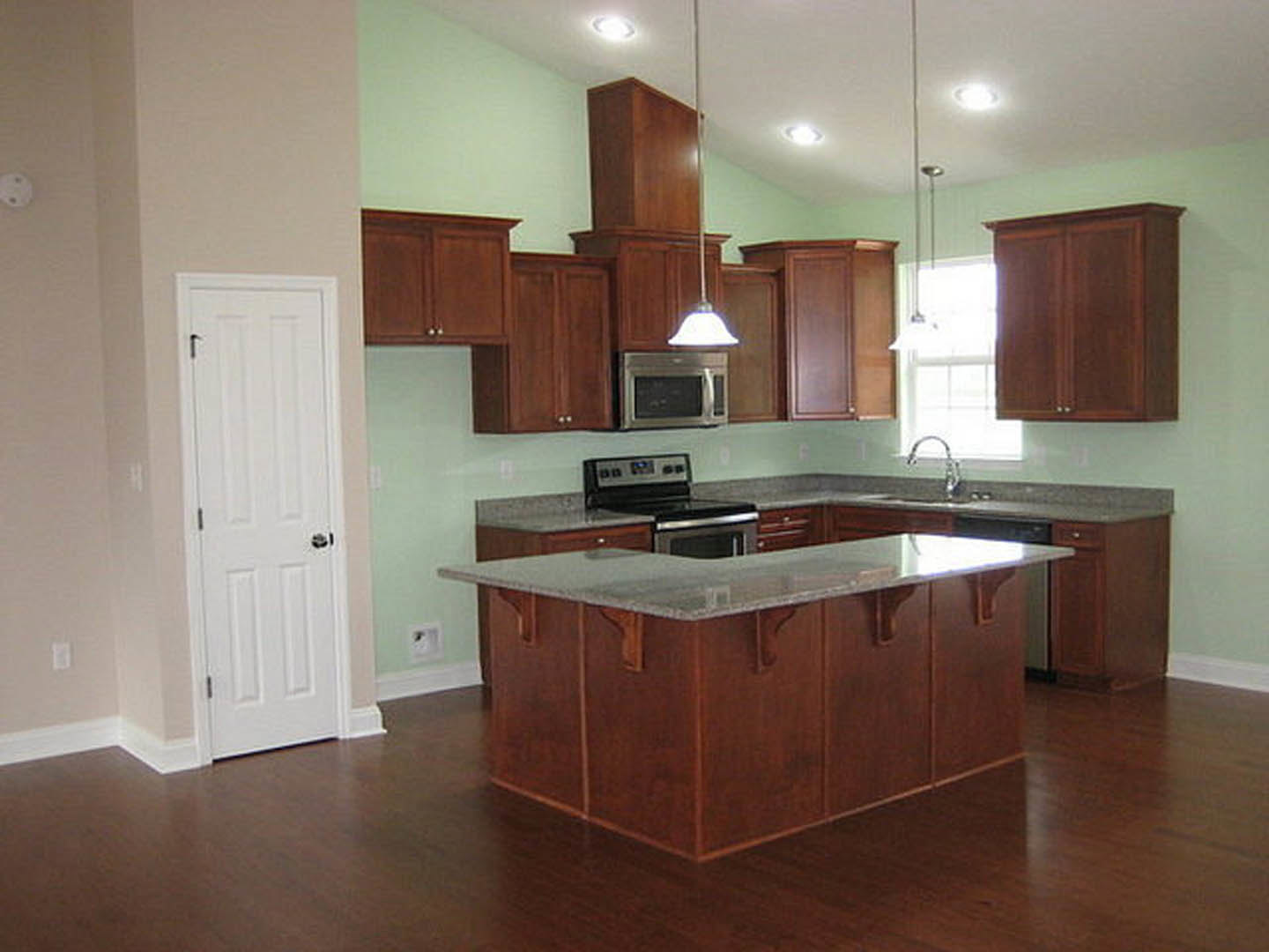 Spacious kitchen featuring a large marble-topped island, white cabinetry, stainless steel appliances, and sunlight streaming through a window onto hardwood floors