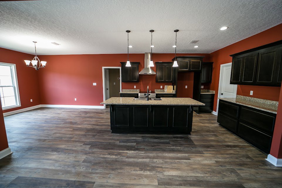 Open kitchen with wood flooring, central island featuring built-in sink, black upper cabinets above red accent wall, modern chandelier, and large window providing natural light