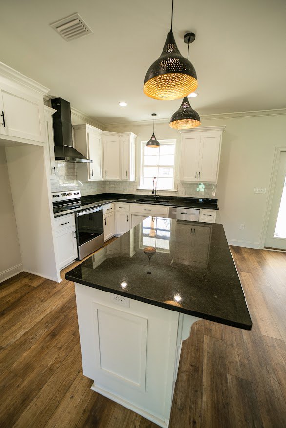 Modern kitchen featuring a black granite island, white cabinetry, gold and black pendant light fixture, stainless steel sink, and large glass window above the counter.