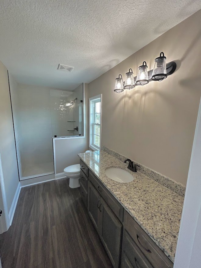 Bathroom featuring a marble countertop vanity, glass-enclosed shower with tile walls, and white toilet adjacent to dark speckled flooring