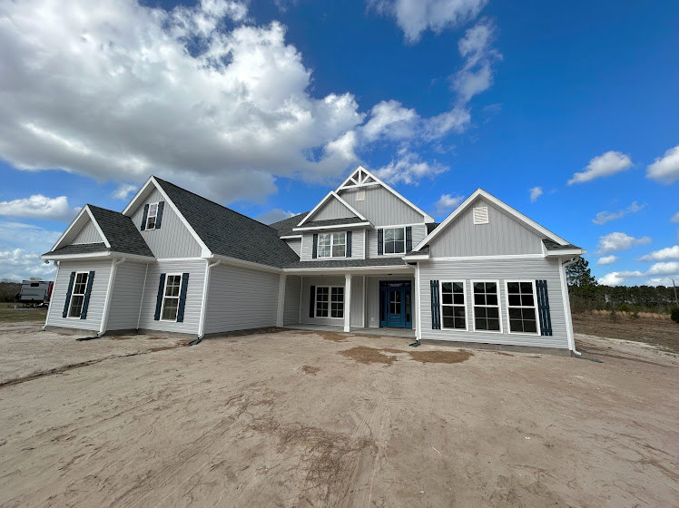 Framed house under construction with exposed plywood walls, blue front door, row of rectangular windows, dirt yard, pitched roof, and partly cloudy blue sky