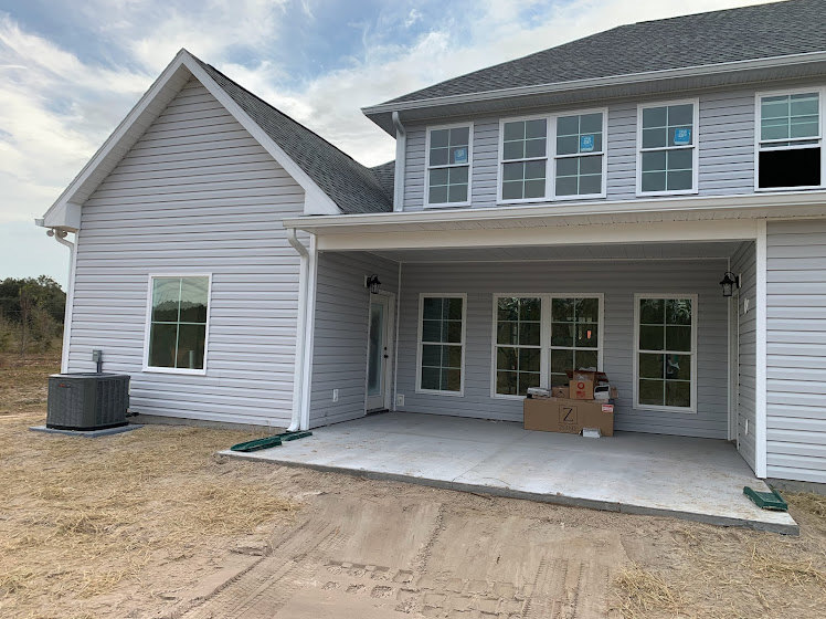 Gray siding two-story home with white trim, covered porch, cardboard boxes stacked near entry, concrete walkway, multiple windows including one with a blue sign, partly cloudy sky