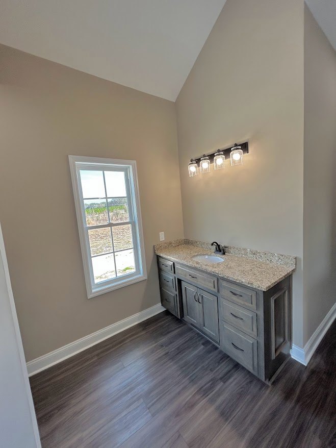 Bathroom with wood flooring, white sink set in cabinetry, wall-mounted lights above mirror, window overlooking grassy field, tile backsplash, drawers beneath countertop