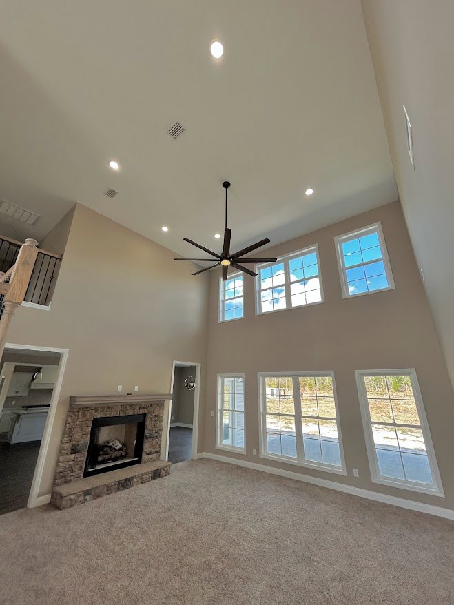 Living room with beige carpet, white plaster walls, black-framed fireplace, ceiling fan with light, large window showing blue sky