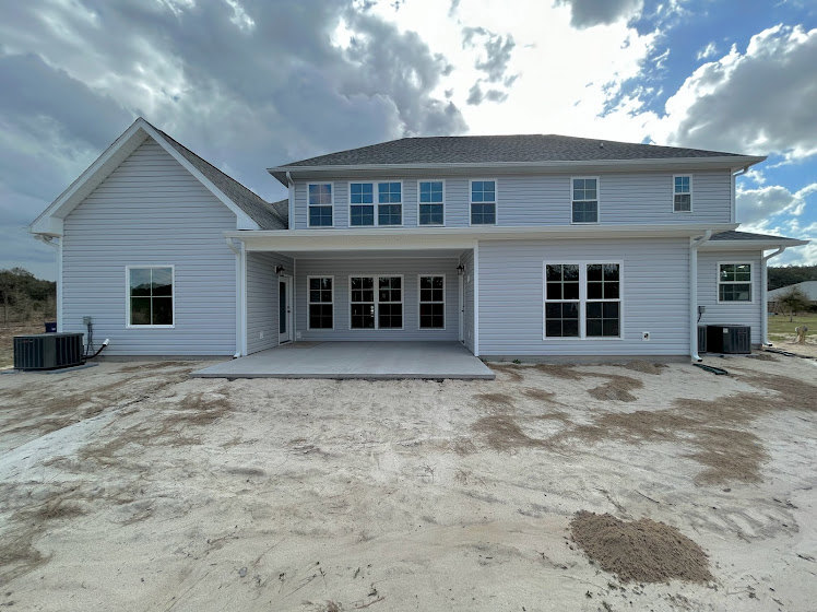 Two-story house under construction with gray siding, multi-pane windows, attached garage, sand-covered driveway, and cloudy sky overhead.