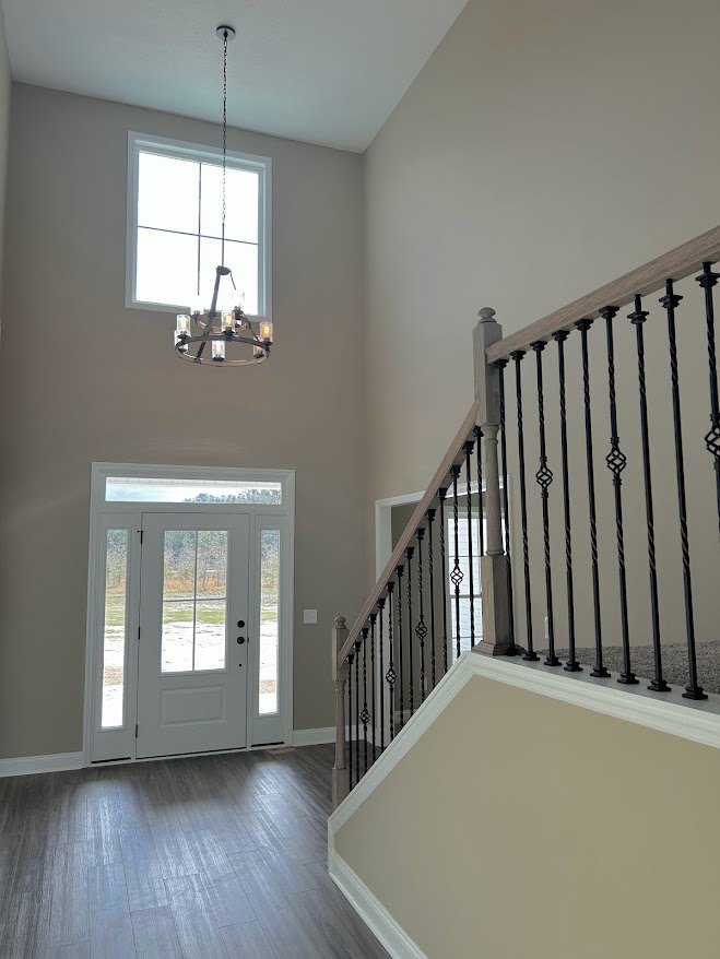 Wooden staircase with black metal railings, white door with glass panes, chandelier hanging near window, light fixture illuminating hardwood floor, white plaster walls.