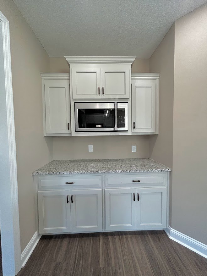White kitchen cabinets with black handles, built-in microwave above countertop, wood flooring, white baseboards, and stainless steel sink.