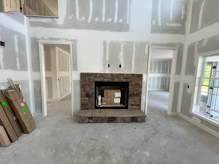 Living room with brick fireplace, white plaster walls, white door, and window with white frame; brown cardboard boxes stacked near hearth.