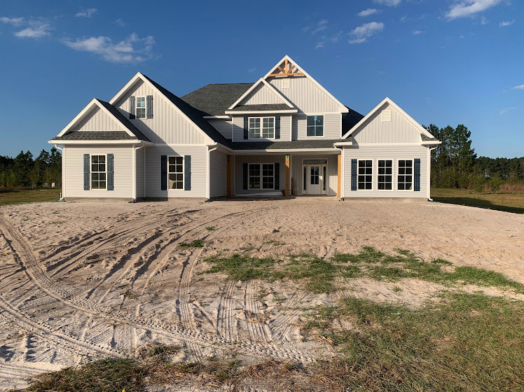 Modern two-story home with black-framed windows, light siding, and dark roof; dirt yard with visible tire tracks in front; cloudy sky overhead