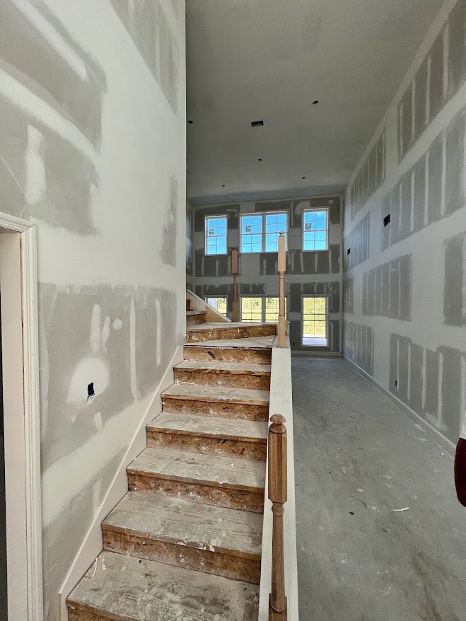 Wood staircase with open risers and white-painted walls, white floor, large window showing blue sky, grey rectangular wall accents, modern interior room.