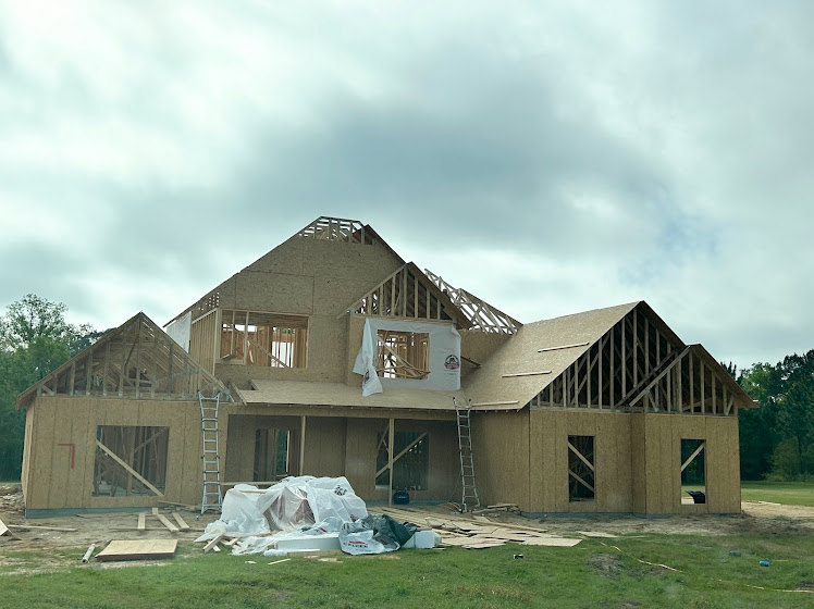 Partially built house with exposed framing, several ladders leaning against exterior walls, surrounded by grassy field and scattered trees under cloudy sky
