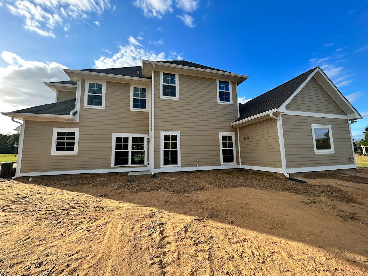 Two-story house with white-framed windows, light-colored siding, and a dirt yard under a partly cloudy blue sky