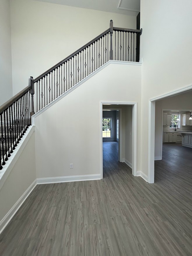 Open wood staircase with black metal railing, light hardwood floors, adjacent kitchen with white cabinets and large window, doorway visible at base of stairs