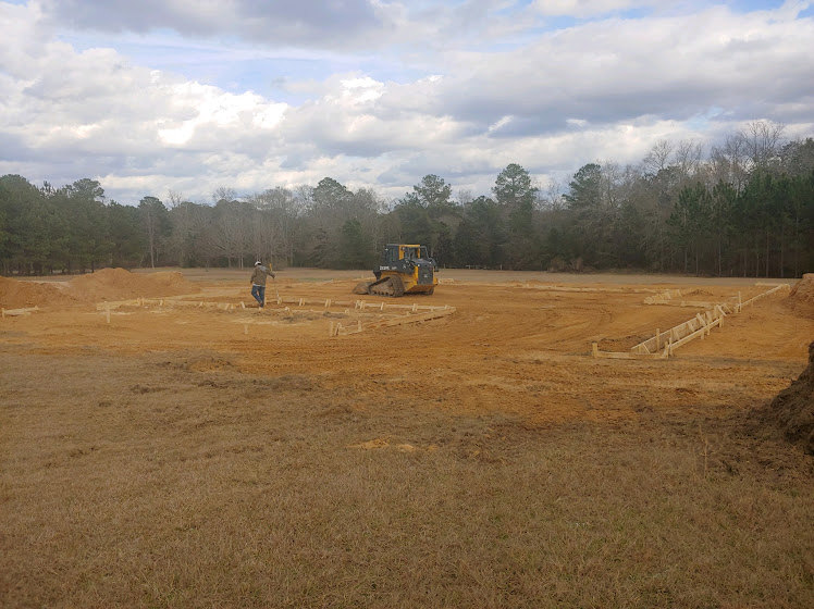 Man standing on grassy lot beside yellow bulldozer, cloudy sky overhead, trees in background