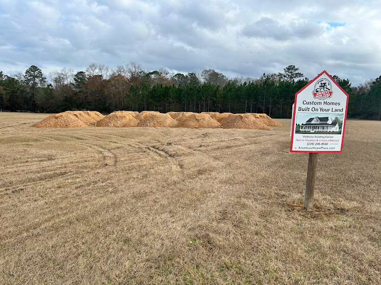 Construction site with a wooden sign, grassy field, piles of dirt, scattered straw, and trees under a cloudy sky