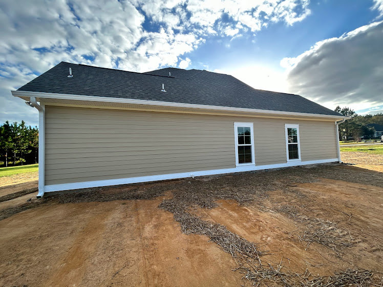 Partially built house with exposed framing, dirt lot in foreground, cloudy sky overhead, visible windows and roof structure