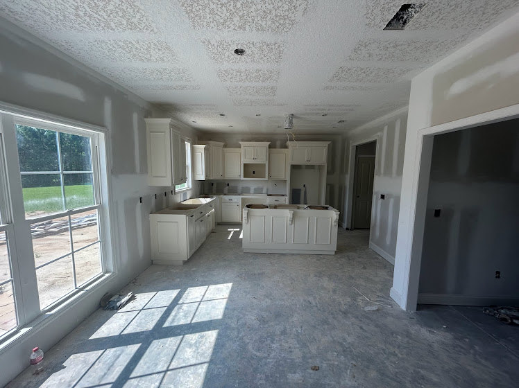 Kitchen under construction featuring unfinished plaster walls, a large window providing daylight, white cabinetry with open shelving, and exposed ceiling and flooring.