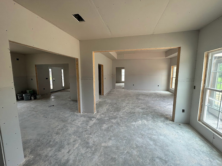 Concrete-floored room with exposed concrete walls, large window, visible pipe, white ceiling featuring a hole, and a white wall displaying a black and white sign