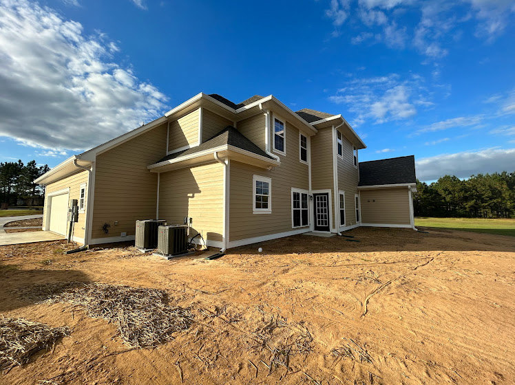 Wood-framed house under construction with exposed beams, unfinished exterior walls, dirt yard, and mature trees in the background