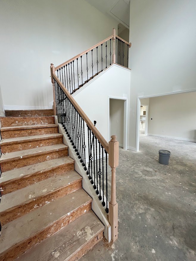 Wood staircase with black metal railing, light switch on hallway wall, grey bucket with lid on concrete floor featuring white spots